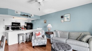 Un salon avec un canapé gris, un fauteuil gris, un pouf gris, et une cuisine blanche équipée d’un four à micro-ondes, d’une cuisinière et d’un réfrigérateur.