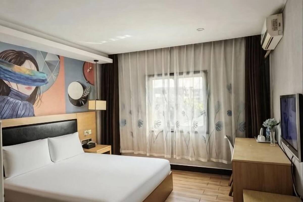 A hotel room with a bed, a desk, a TV, a painting on the wall, and a window with curtains.