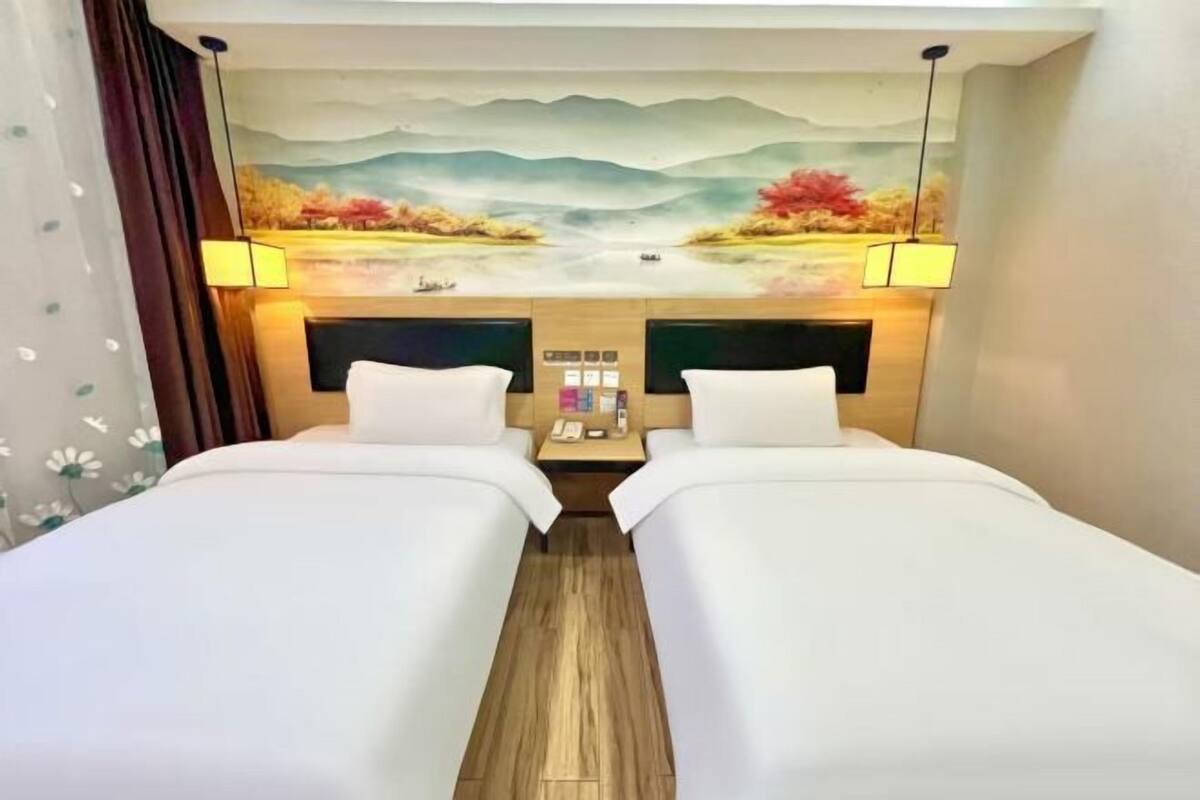 A hotel room with two beds, a wooden floor, a painting of a landscape on the wall, and a nightstand with a lamp.