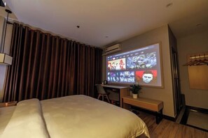 Ein Hotelzimmer mit einem großen Bett, einem Schreibtisch mit Stuhl, einem kleinen Tisch mit Pflanze und einem an der Wand montierten Projektor, der Bilder zeigt.