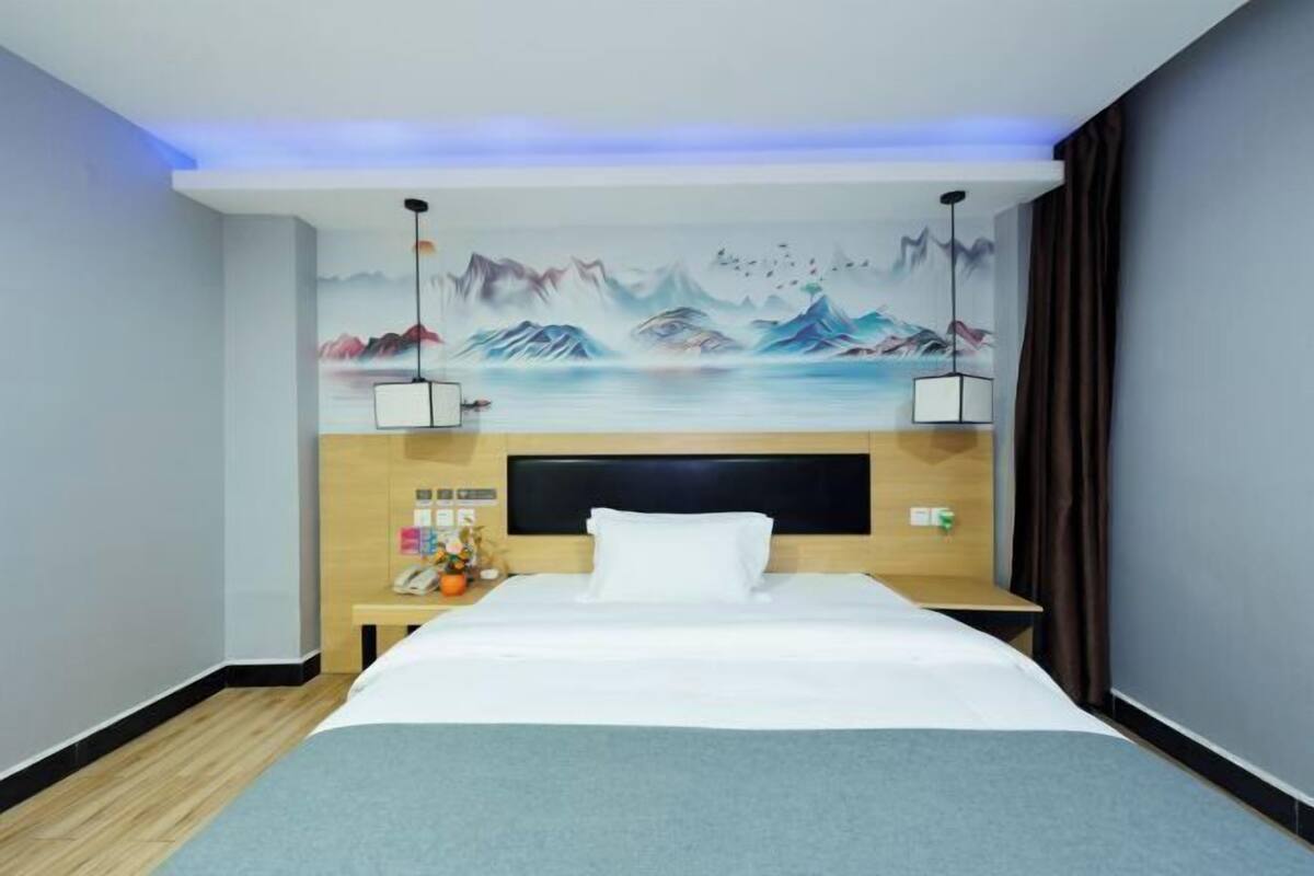 A hotel room with a bed, a nightstand, a wall-mounted phone, a painting of mountains, and a ceiling light fixture.