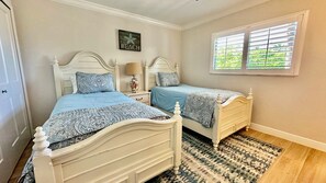 Un dormitorio con dos camas, una ventana con persianas, un adorno en la pared y una alfombra estampada.
