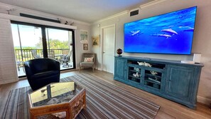 A living room with a large flat-screen TV, a glass-top coffee table, a blue cabinet with decorative items, a black chair, and a view through a sliding glass door.