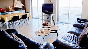 A modern living room with a dining area, a flat-screen TV, and large windows offering a view. - Apartment in Nieuwpoort With Ocean Views (Nieuwpoort)