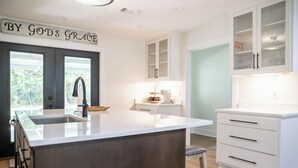 A modern kitchen with a central island, white cabinetry, and a sign above the door. - With GOLF CART! 1 mile ride to the beach & Downtown Ocean Springs! Clark Cottage (Ocean Springs)