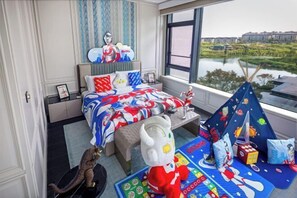 A bedroom with a large window, a bed with a superhero-themed bedspread, a small table, and a teepee with toys.