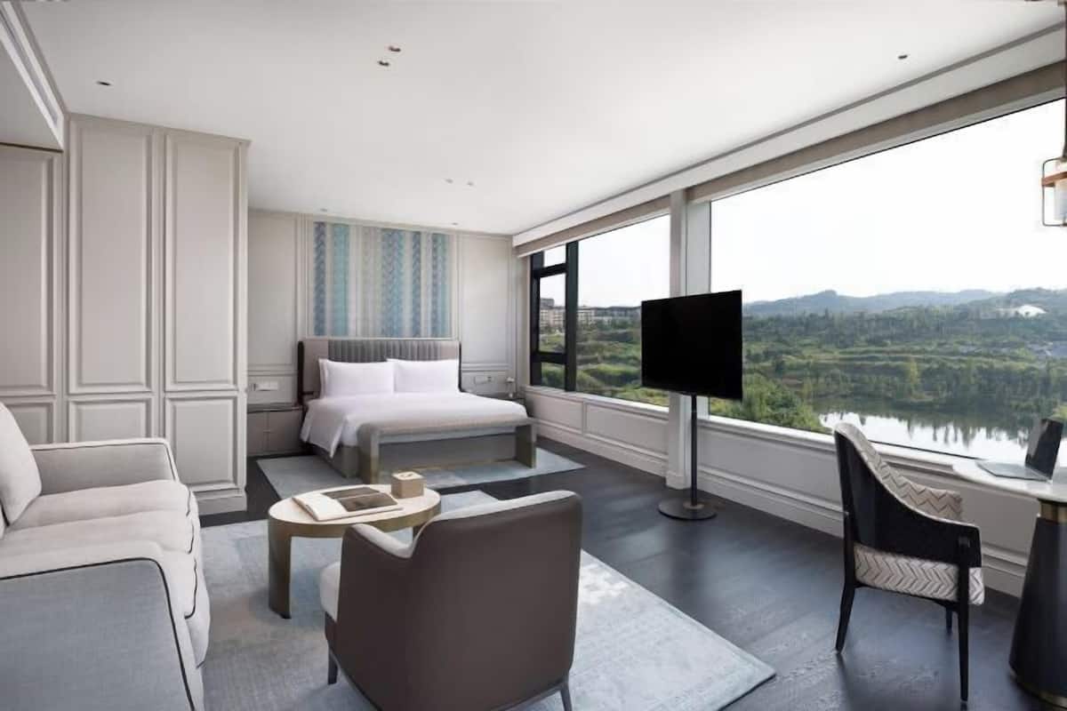 A modern hotel room with a large bed, a TV, a sofa, and a view of a lake.