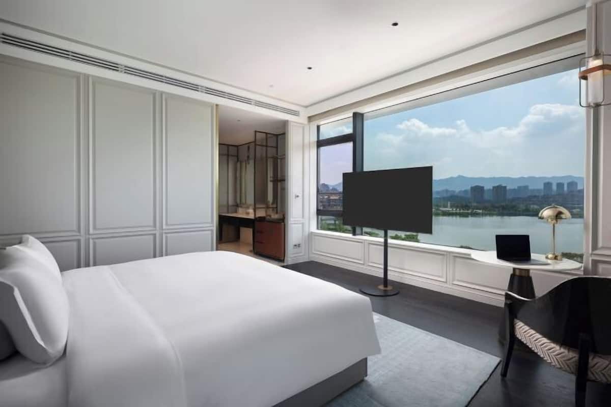A hotel room with a large bed, a TV, a desk, and a view of a lake and cityscape.