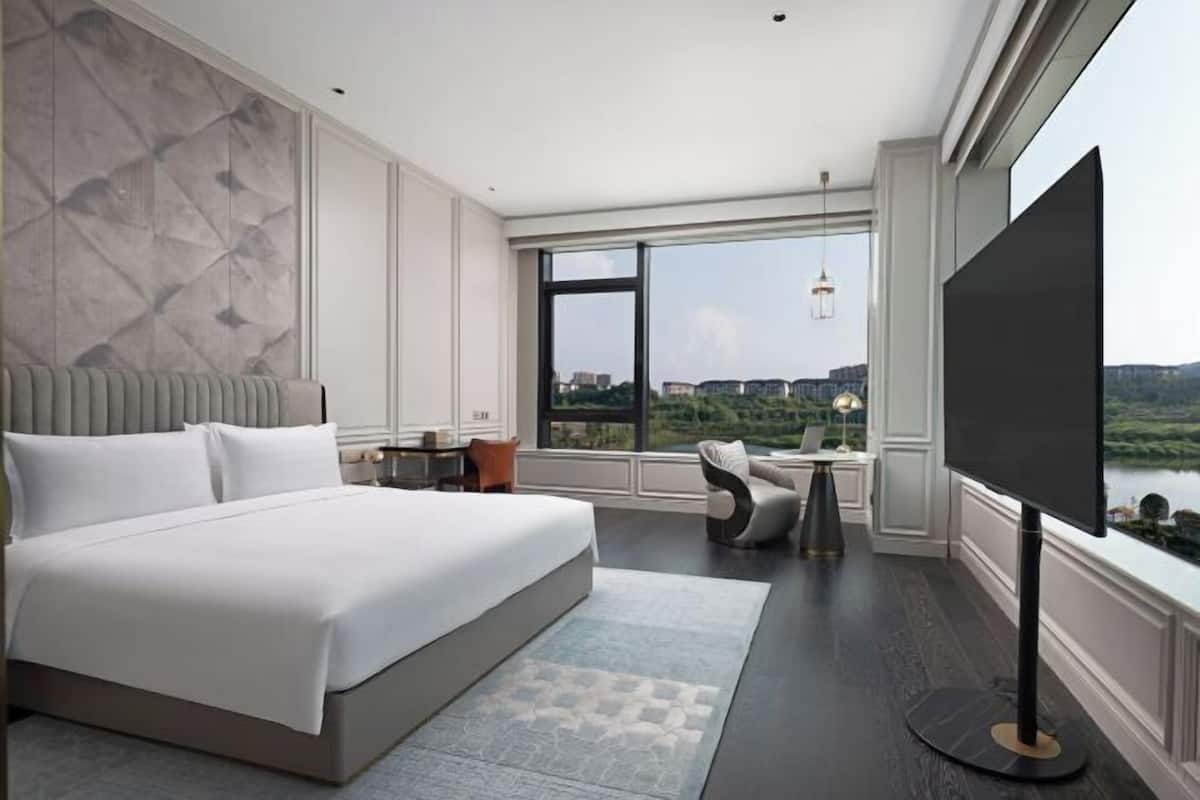 A modern hotel room with a large bed, a desk, a chair, a TV, and a view of the city.