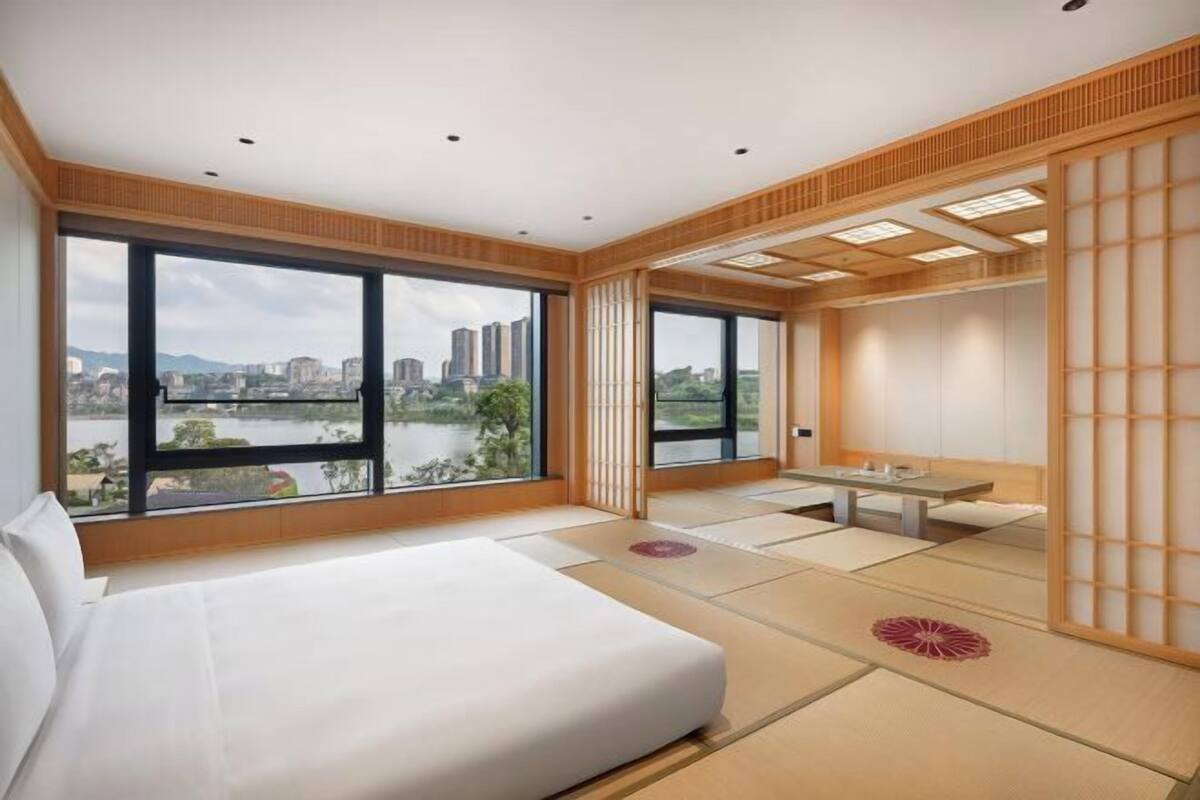 A hotel room with a large bed, a dining table, tatami flooring, a view of a cityscape, and a traditional sliding door.