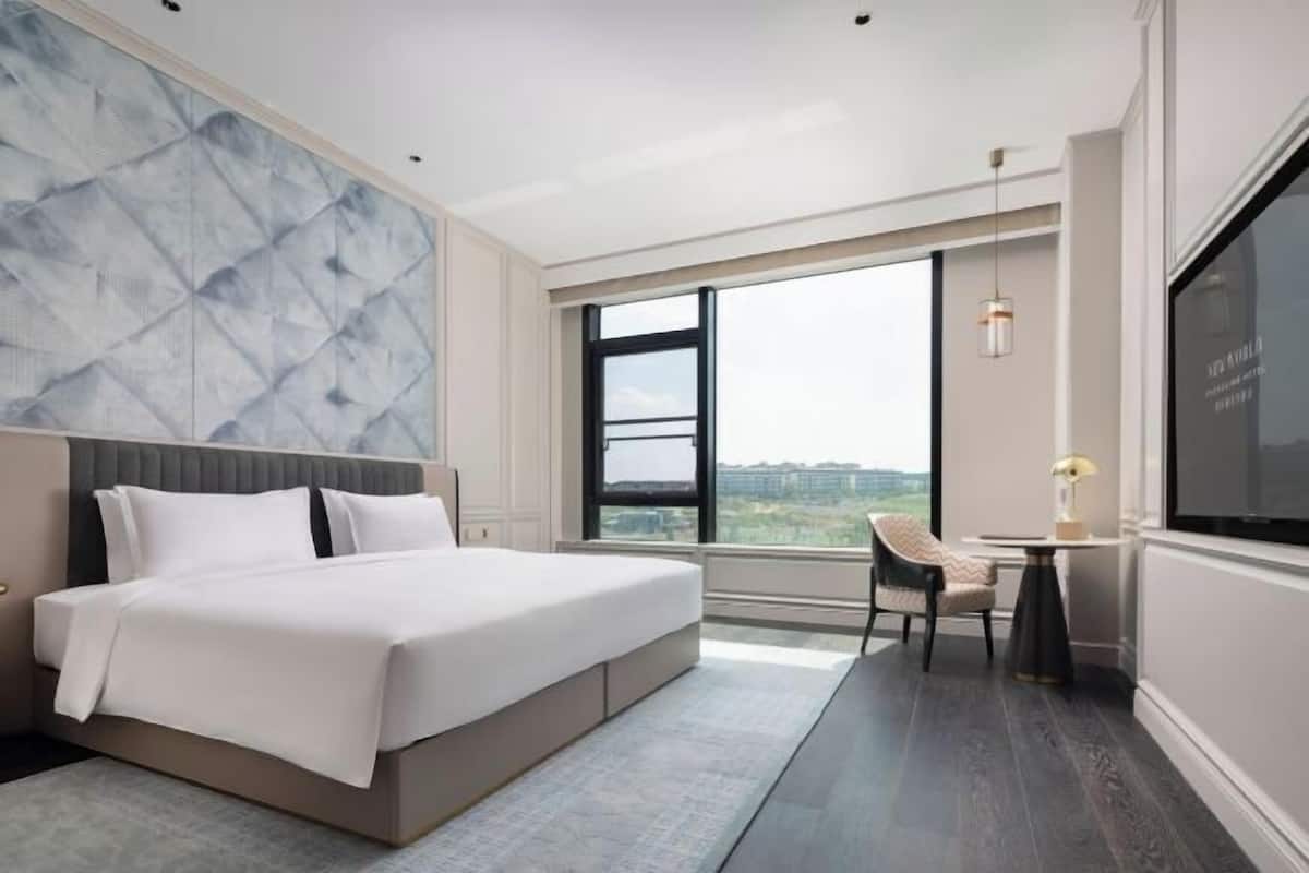 A modern hotel room with a large bed, a view through a large window, and a small table with a chair.