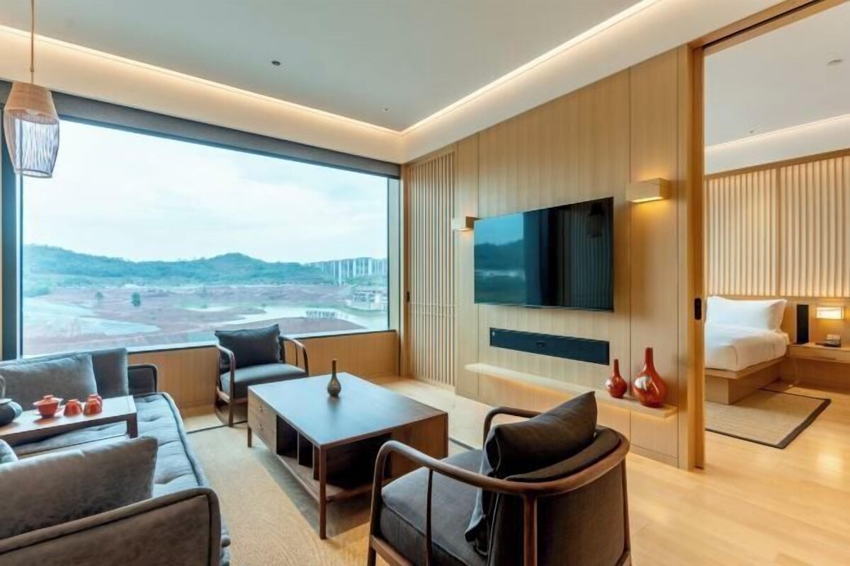 A modern hotel room with a large window offering a view of the beach, a flat-screen TV mounted on the wall, a sofa, a dining table, and a bed with a nightstand.