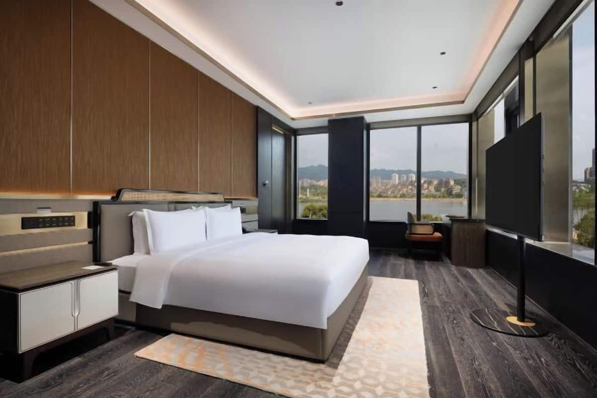 A modern bedroom with a large bed, bedside tables, a TV, and a city view.