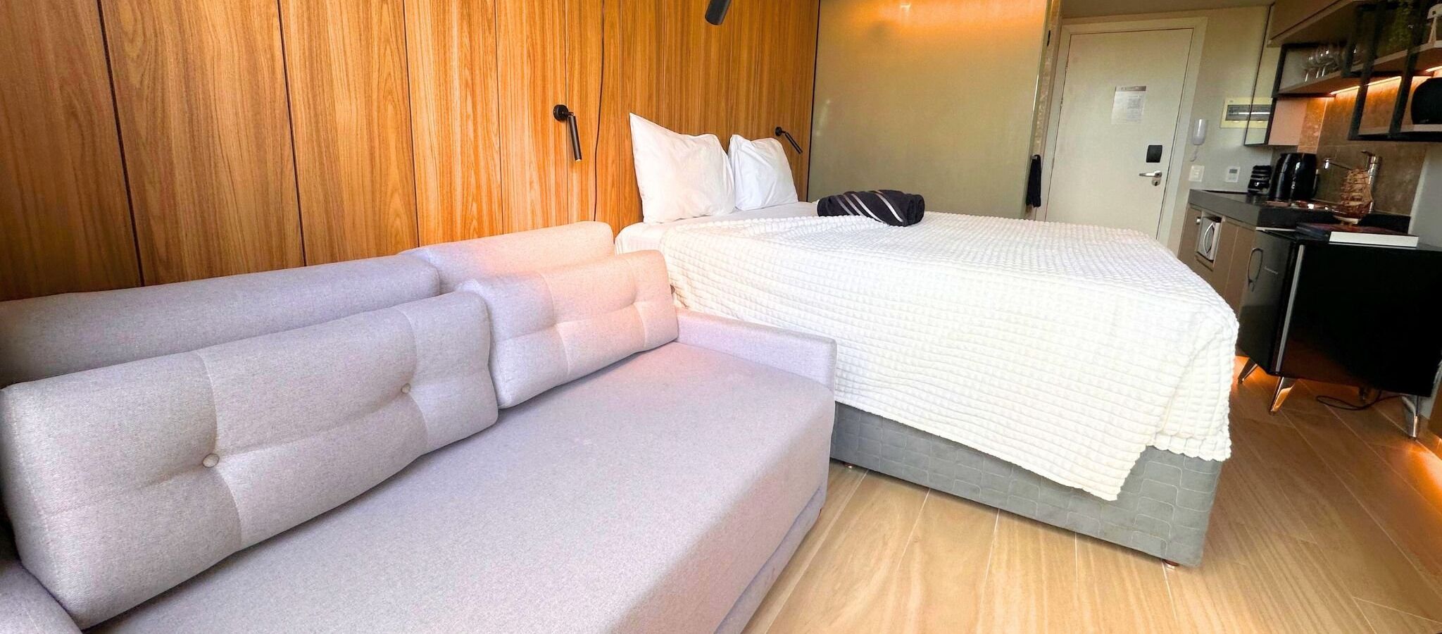 A modern hotel room with a sofa, a bed with a white quilt, wooden paneling on the walls, and a kitchenette area.