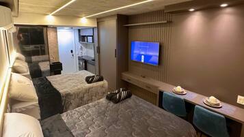 Quarto de hotel compacto com duas camas, televisão, mesa pequena para refeições e vista para o corredor.