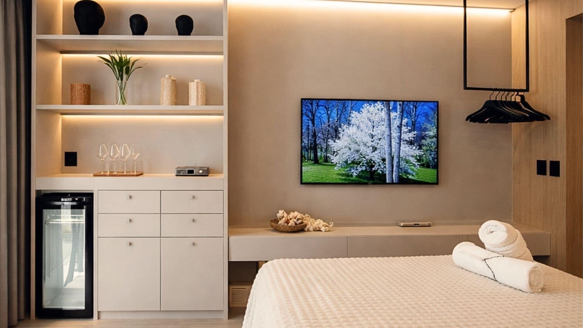 A modern hotel room with a bed, a TV, a minibar, a built-in oven, and a wall-mounted shelf with decorative items.