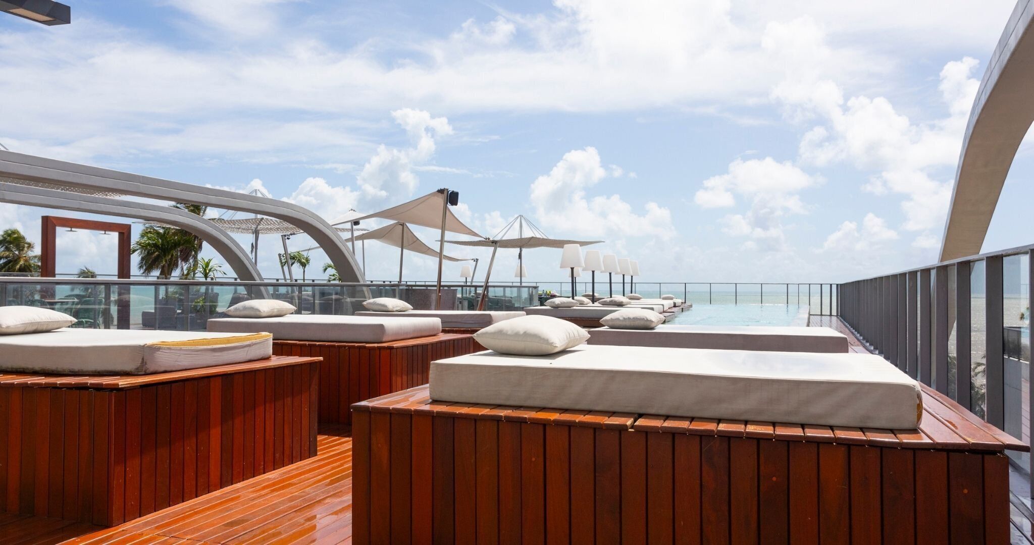A deck with wooden benches and cushions, a glass railing, and a view of the sea.