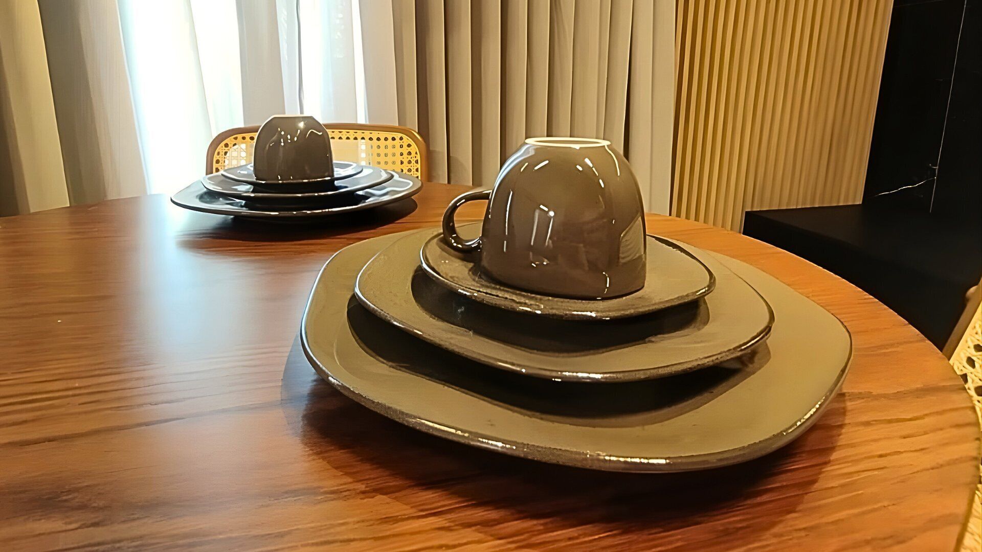 A round dining table with two sets of plates and cups, a chair, and a window with blinds.