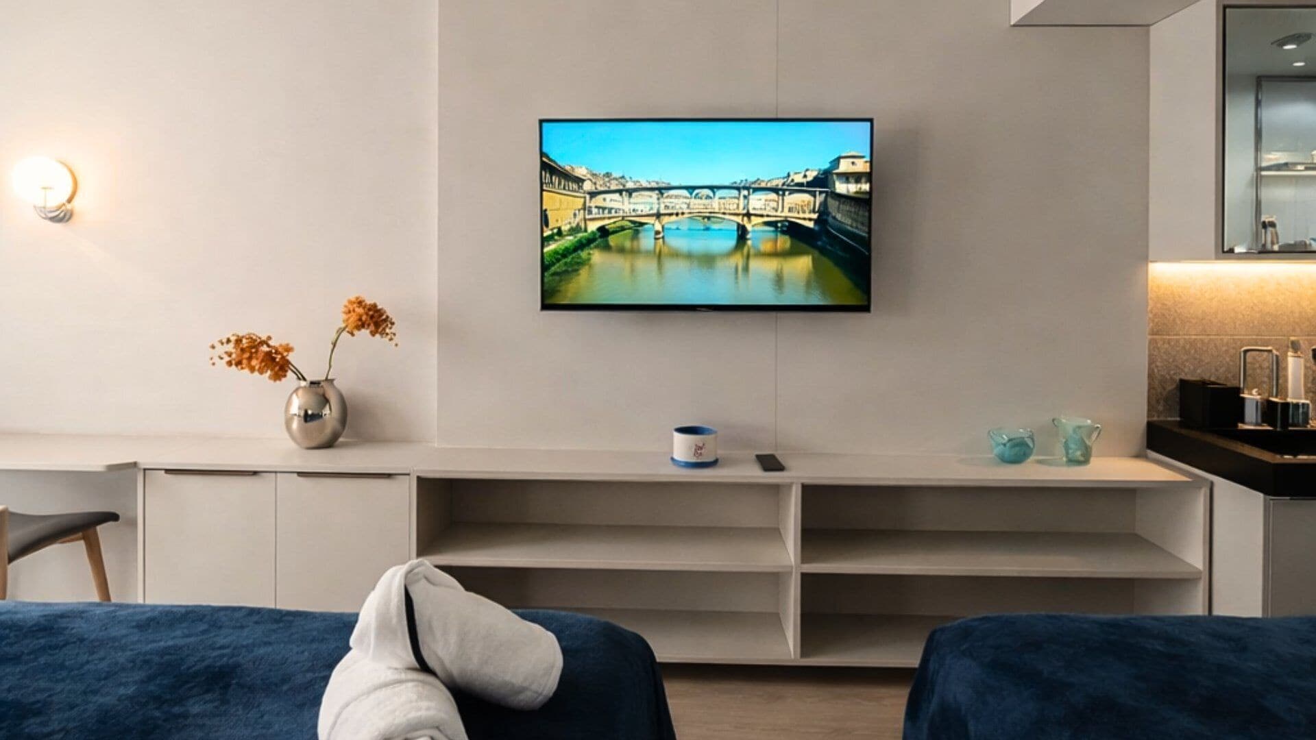 Una habitación de hotel moderna con un televisor de pantalla plana en la pared, una mesita de noche con un jarrón de flores y una cama con cubierta azul.
