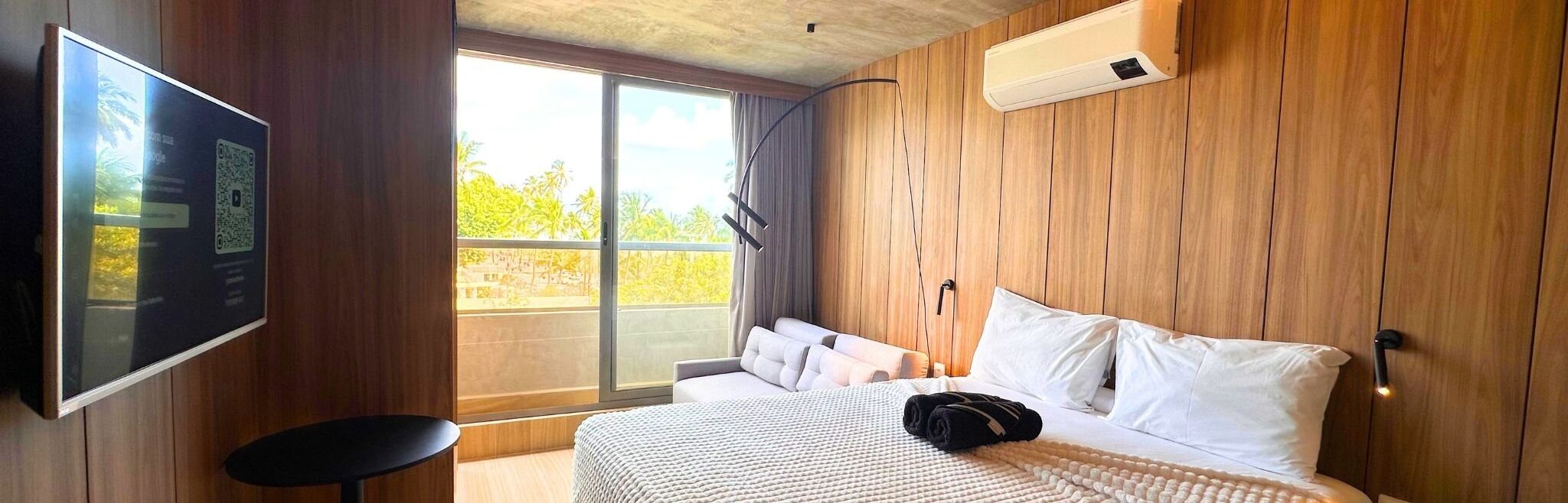 Une chambre d’hôtel moderne dotée d’un grand lit, d’une télévision à écran plat, d’une petite table et d’un balcon offrant une vue sur la verdure.