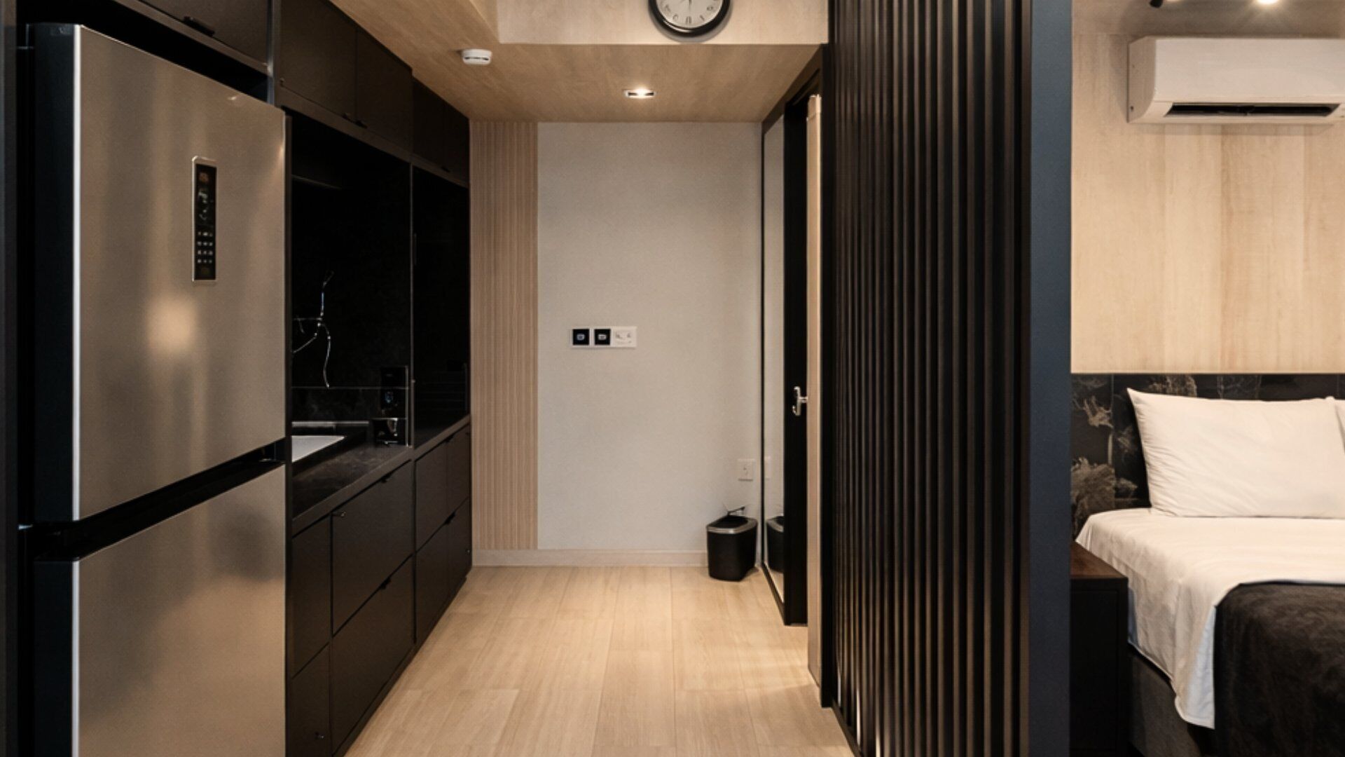 Une chambre d’hôtel compacte dotée d’une kitchenette, d’un lit avec du linge de maison blanc, d’un plancher en bois et d’un climatiseur fixé au mur.