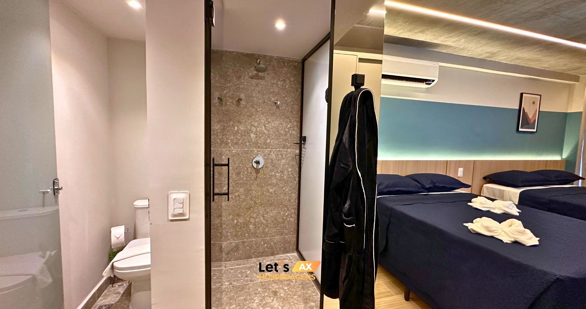 Quarto de hotel com chuveiro, vaso sanitário, cama com duas almofadas e mesa de cabeceira.