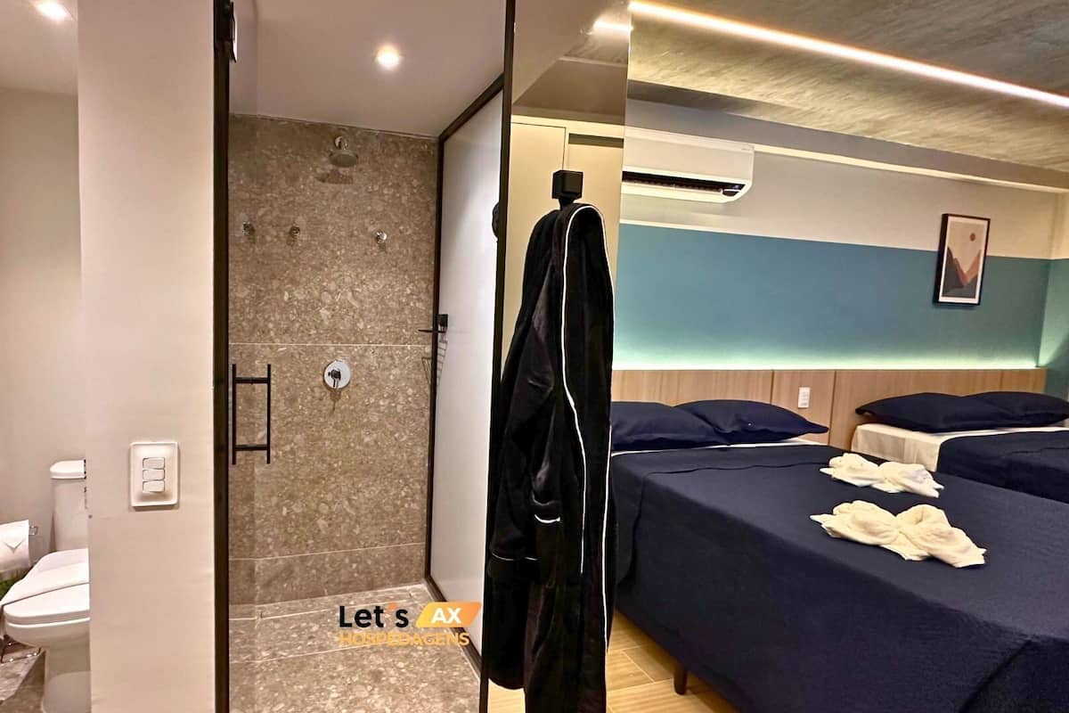 A hotel room with a shower, a toilet, a bed with two pillows, and a bedside table.