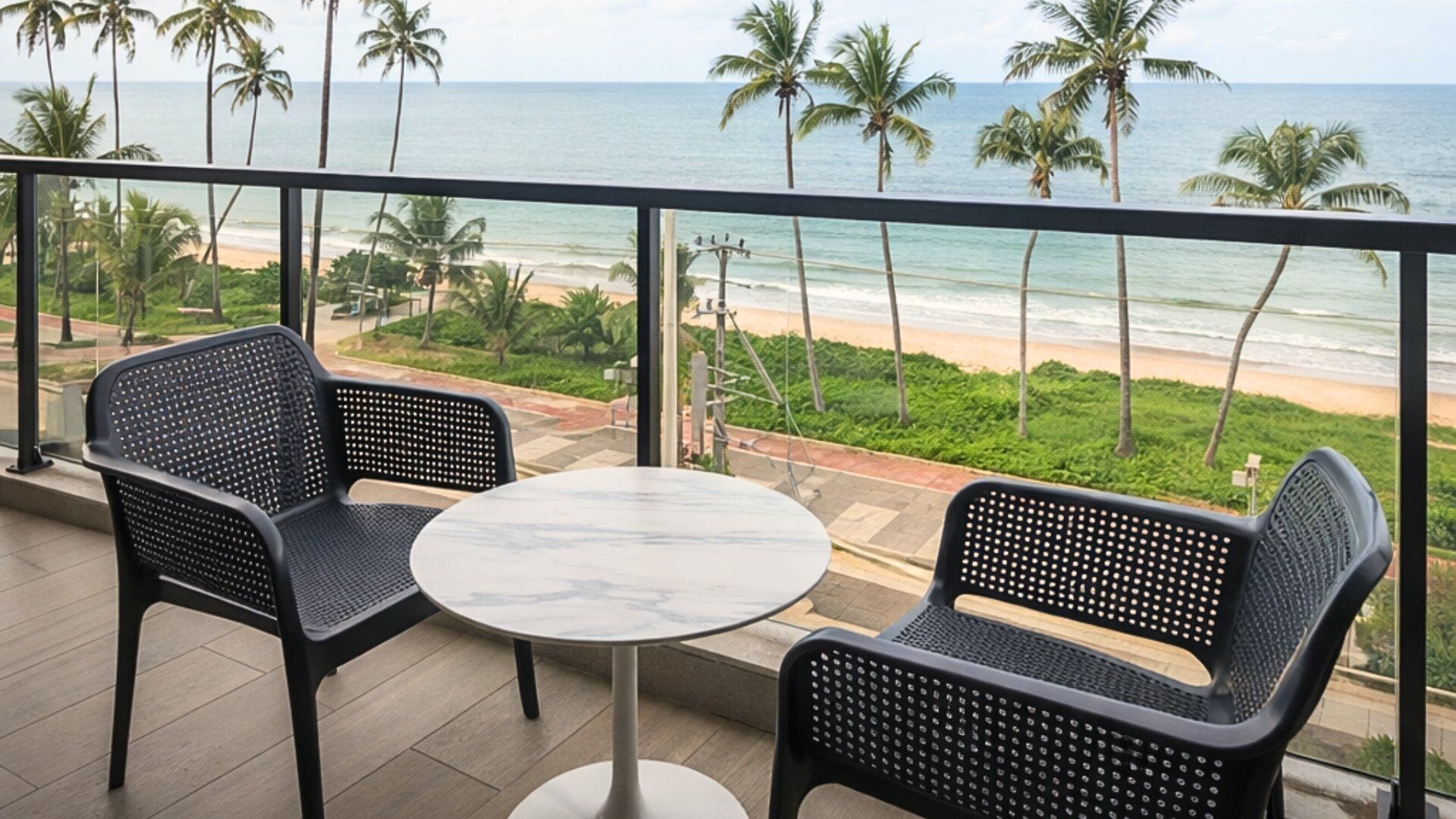 A balcony with two chairs and a table, overlooking a beach with palm trees and the ocean.