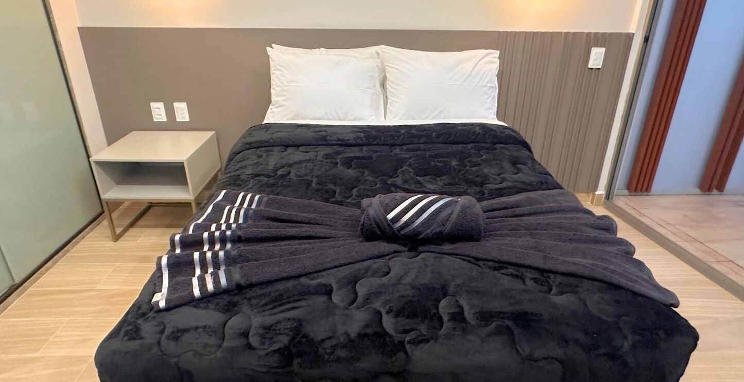 A neatly made bed with a dark cover, a white pillow, and a striped towel.