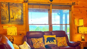 Ein gemütliches Blockhaus-Interieur mit Ledersofa, gemusterten Kissen und Blick auf Grünfläche.