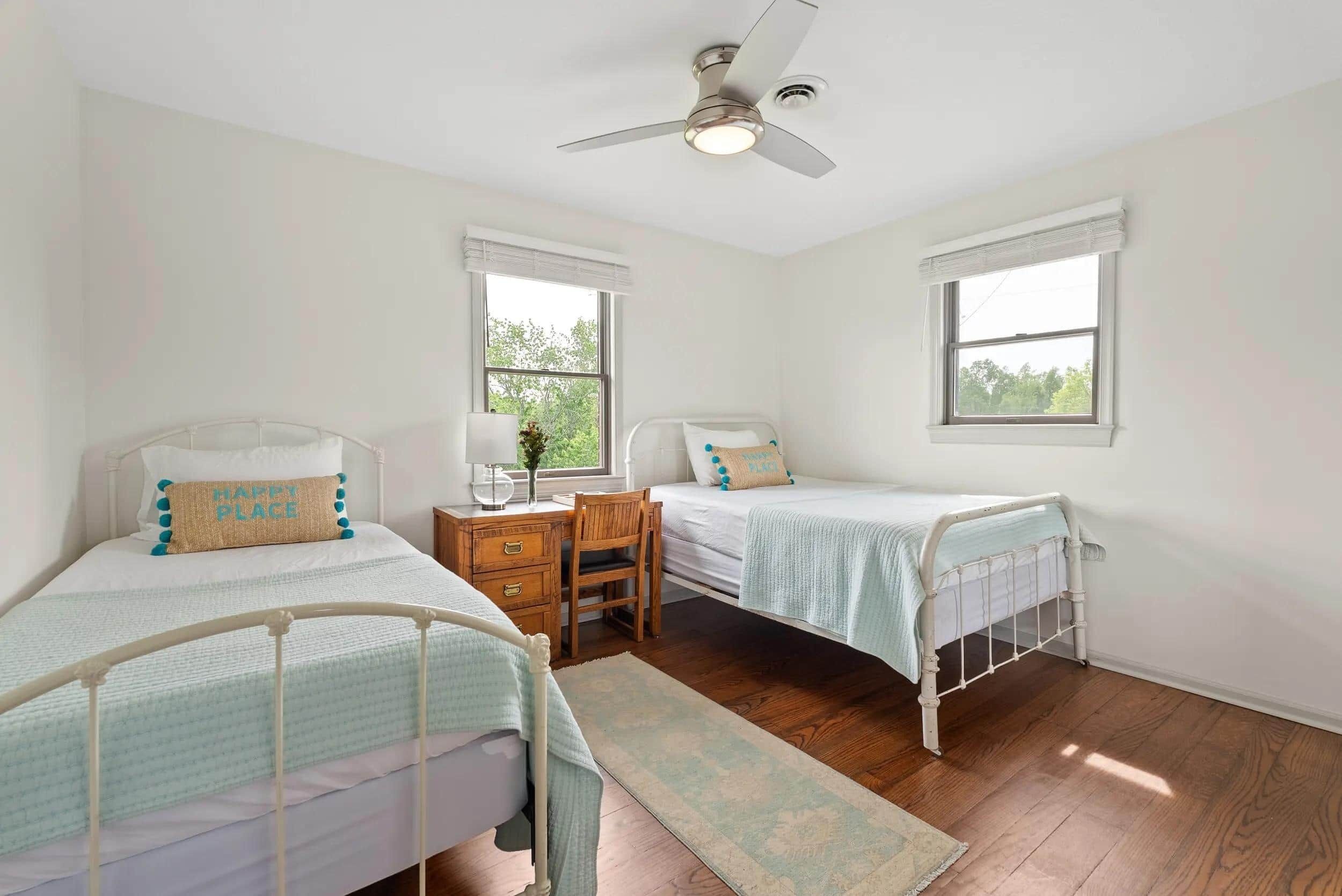 Un dormitorio con dos camas individuales, un escritorio, una silla, un ventilador de techo y dos ventanas.