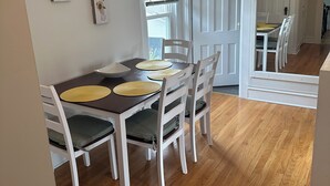A dining area with a wooden table and white chairs, a window with blinds, and two wall-mounted paintings.