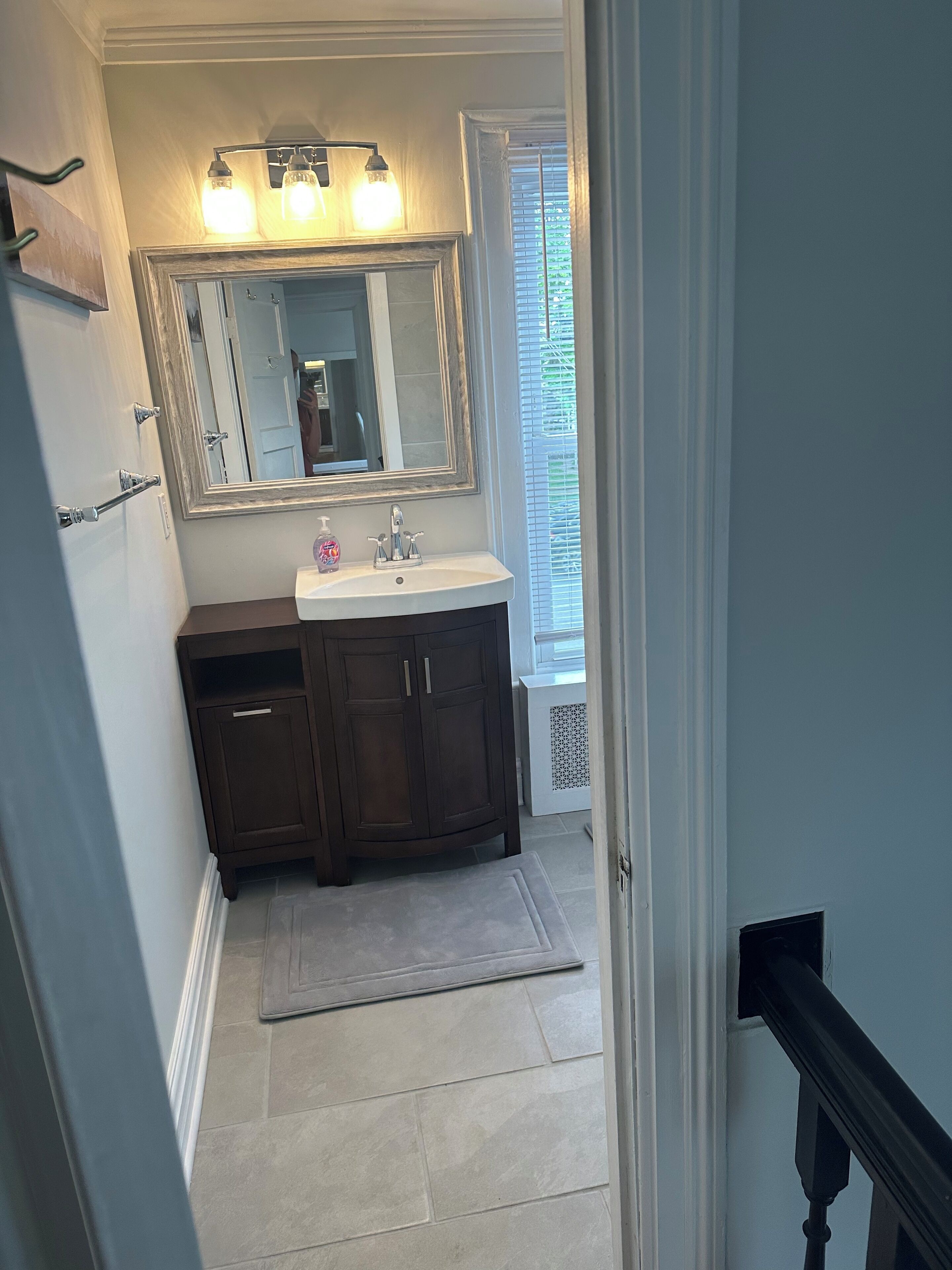A bathroom with a dark wooden vanity, a white sink, and a large mirror above it. There is a window with blinds to the right and a door leading to another room.