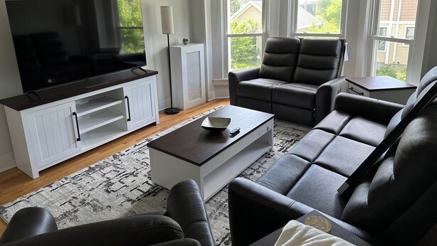 A living room with a flat-screen TV, a white entertainment unit, a grey sofa, a coffee table, and a window with blinds.