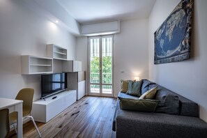 Una sala de estar moderna con un televisor de pantalla plana, un sofá gris con cojines, un escritorio blanco y una estantería.