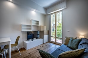 A modern living room with a flat-screen TV, a white entertainment unit, a sofa with cushions, a chair, and a large window with a view of greenery.