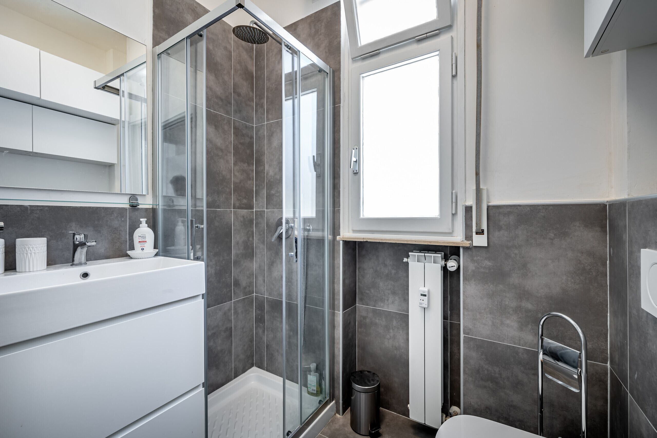 A modern bathroom with a glass-enclosed shower, a white vanity with a mirror, and a window providing natural light.