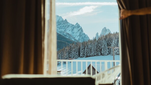 Vista a través de una ventana con cortinas, mostrando árboles y montañas cubiertas de nieve.
