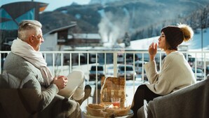 Una pareja sentada en un balcón con vista a la montaña, una cesta de mimbre con comida y un vaso de bebida sobre la mesa.