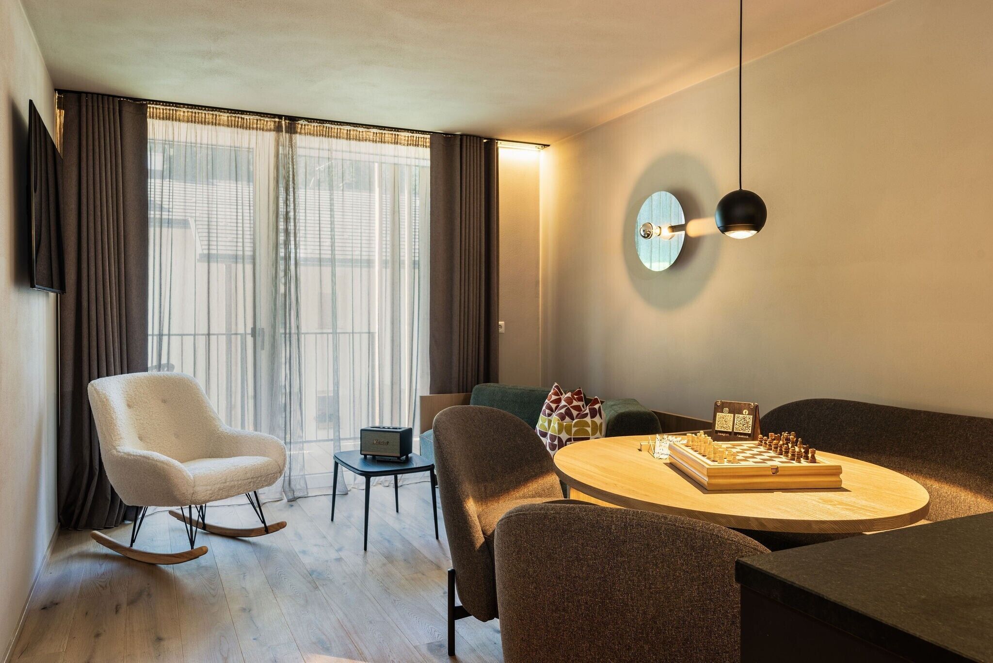 Un salon moderne avec une table ronde en bois, un fauteuil à bascule blanc, un canapé et un téléviseur fixé au mur.