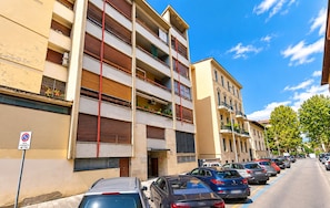 A row of multi-story residential buildings with balconies, parked cars, and a clear blue sky.