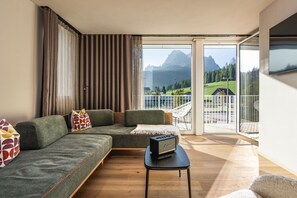 A modern living room with a sectional sofa, a small table with a speaker, and a balcony with a view of mountains. - Ariane's Guesthouse – Relax: Apartment with View of the Sesto Dolomites (Sesto)