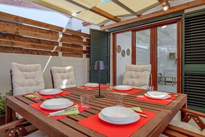 Een terras met een houten tafel, rode placemats, witte borden en stoelen met kussens.