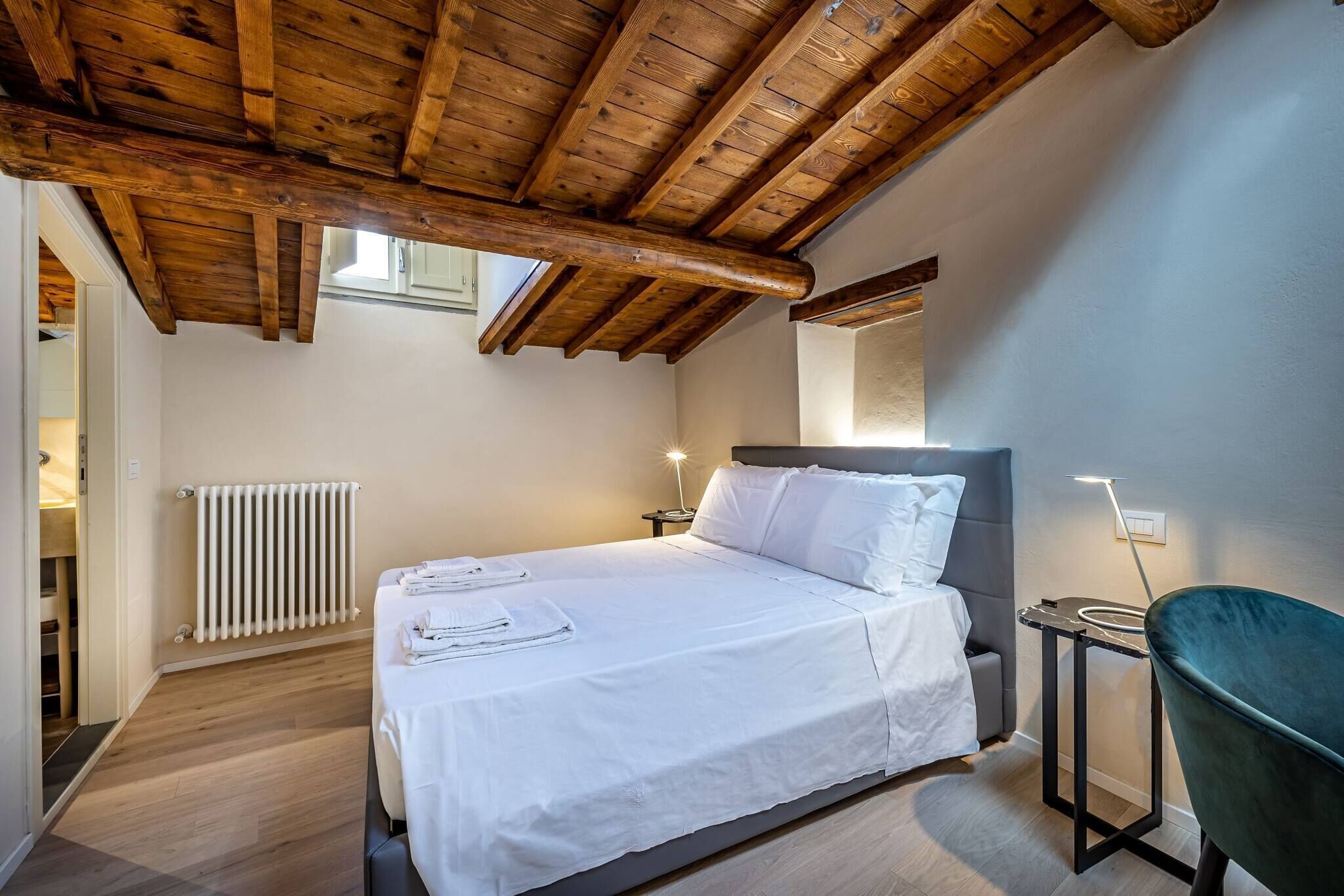 Une chambre avec un plafond en bois, un lit recouvert de draps blancs, une table de chevet, une chaise et un radiateur.