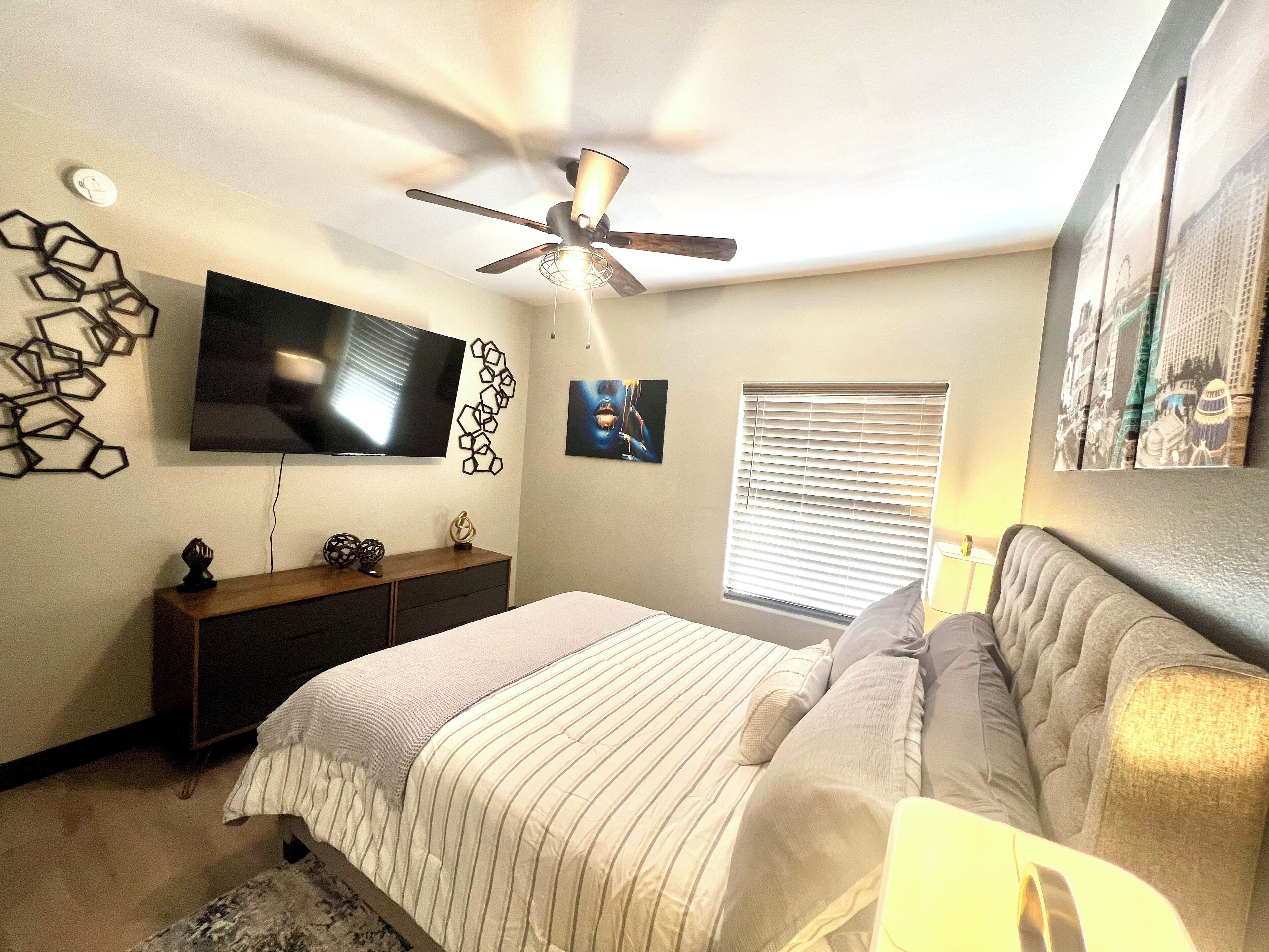 A bedroom with a bed, a ceiling fan, a TV, a dresser, and a window with blinds.