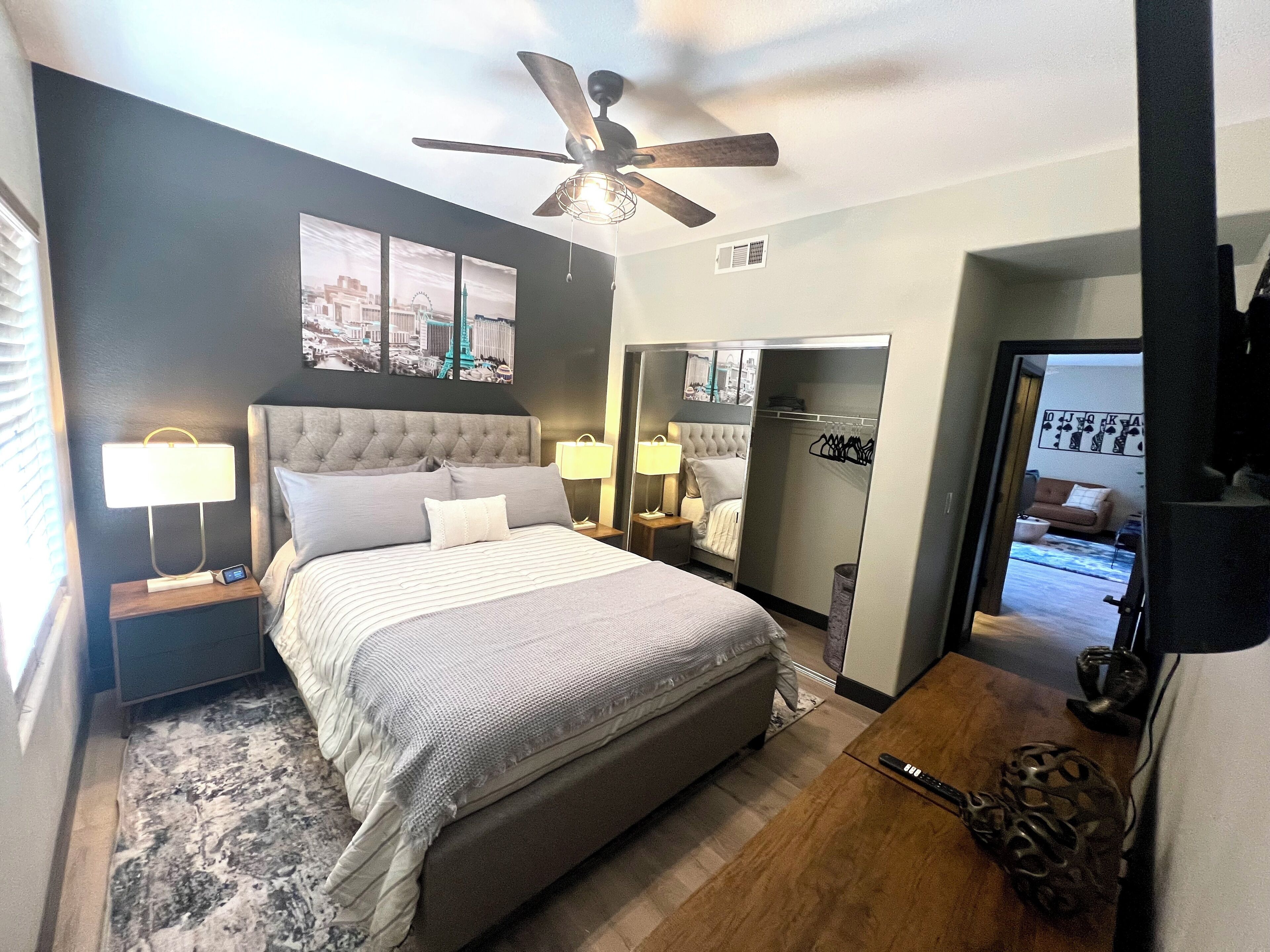 A bedroom with a large bed, two bedside tables with lamps, a ceiling fan, a wardrobe, and a view of the cityscape through the window.