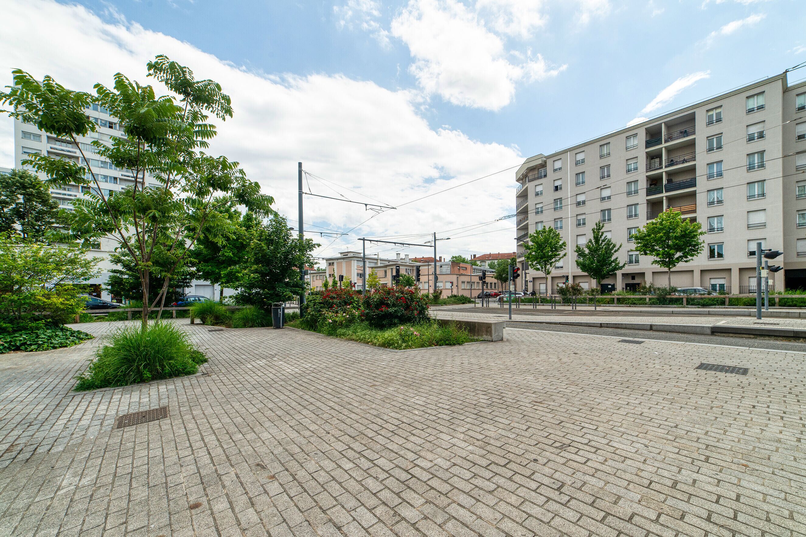 A paved pedestrian area with greenery and a multi-story building in the background.