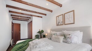 Een slaapkamer met een bed, een groene deken, witte handdoeken, houten balken, een nachtkastje, een lamp, een plant en twee ingelijste foto's aan de muur.