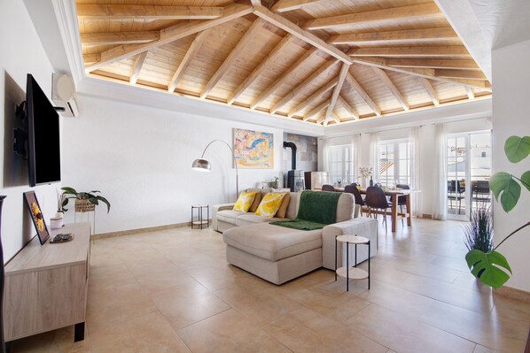 A spacious living area with a high wooden ceiling, a sofa with cushions, a dining table, and a TV mounted on the wall.