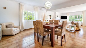 Un comedor con mesa de madera y sillas tejidas, un sofá y un televisor en una habitación luminosa con ventanas amplias.
